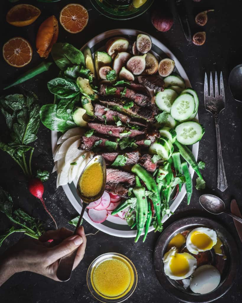 spoon pouring tangerine turmeric dressing on steak salad with avocados cucumbers figs radishes jicama